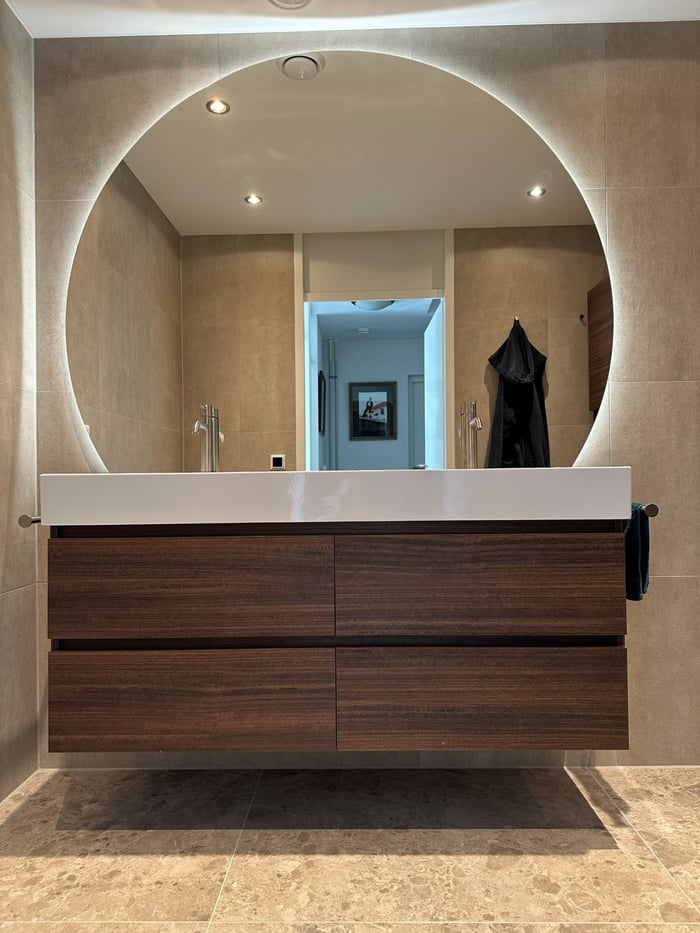Zwevend walnoten wastafelmeubel met brede witte wastafel en grote ronde spiegel met indirecte verlichting, in een badkamer met beige tegels op wand en vloer.