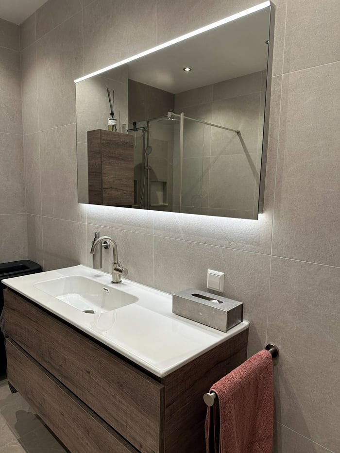 Badkamer met zwevend wastafelmeubel in grijs eiken, witte wastafel en brede spiegel met indirecte verlichting, tegen beige steenlook-tegels.