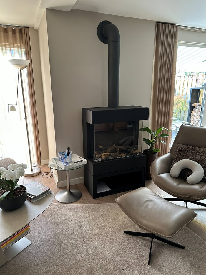 Woonkamer met een vrijstaande zwarte houtkachel met glas aan drie zijden, een leren loungestoel met hocker en een houten vloer.