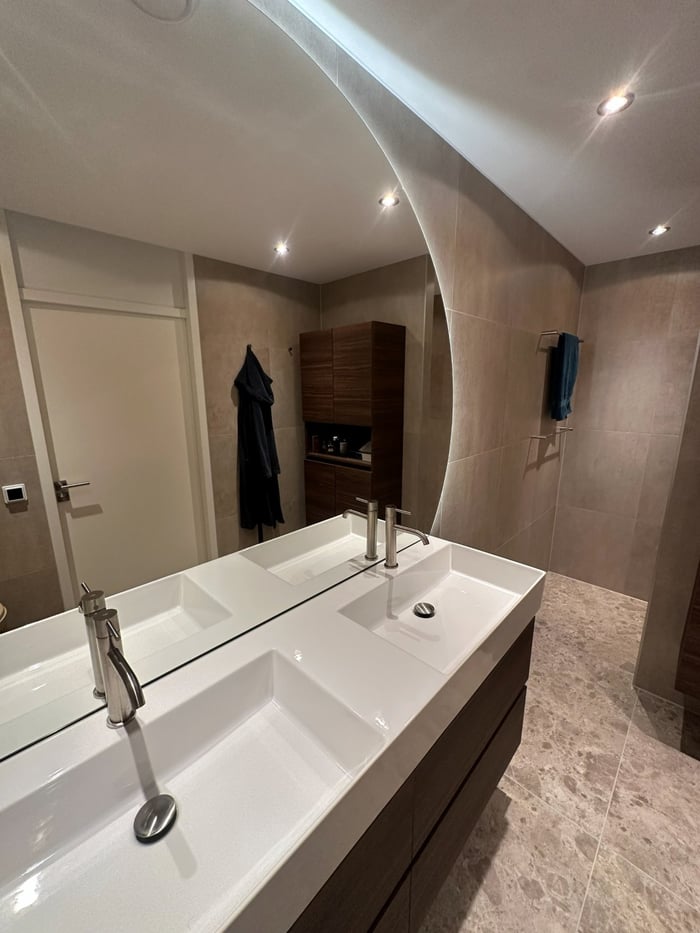 Overzicht van de badkamer met dubbele wastafel, ronde verlichte spiegel en doorkijk naar de inloopdouche en walnoten kolomkast.