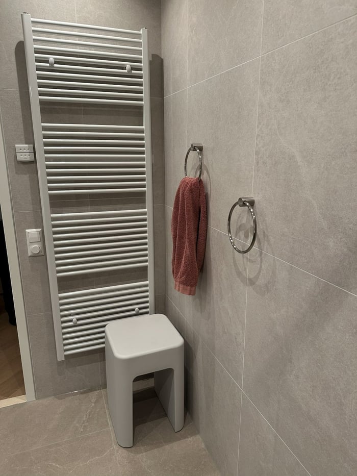 Witte handdoekradiator met handdoekring en een kruk, tegen beige steenlook-tegels.