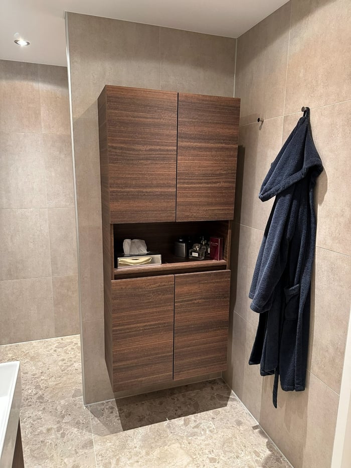 Walnoten kolomkast met open nis tegen een wand van beige tegels, met een badjas aan een rvs haak.