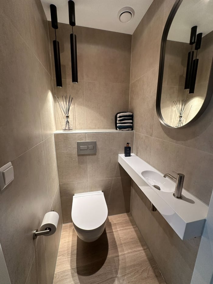 Toiletruimte op de benedenverdieping met hangend toilet, zwevende maatwerk fontein, ovale spiegel en zwarte designverlichting.