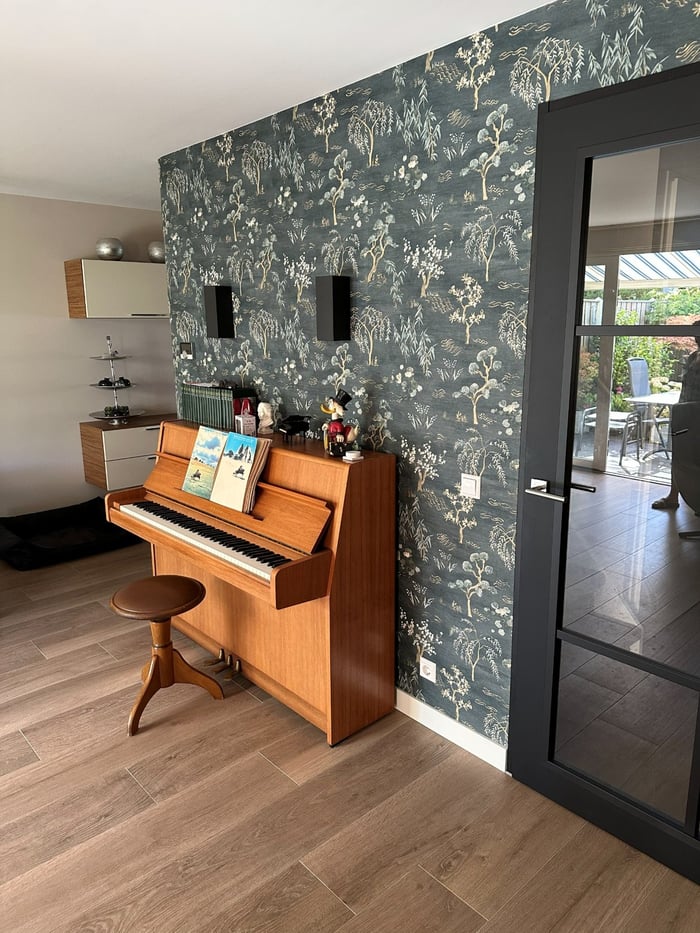 Woonkamer met een piano tegen een donkere wand met botanisch behang, naast een zwarte stalen deur met glas en zicht op de tuin, op een houten vloer.